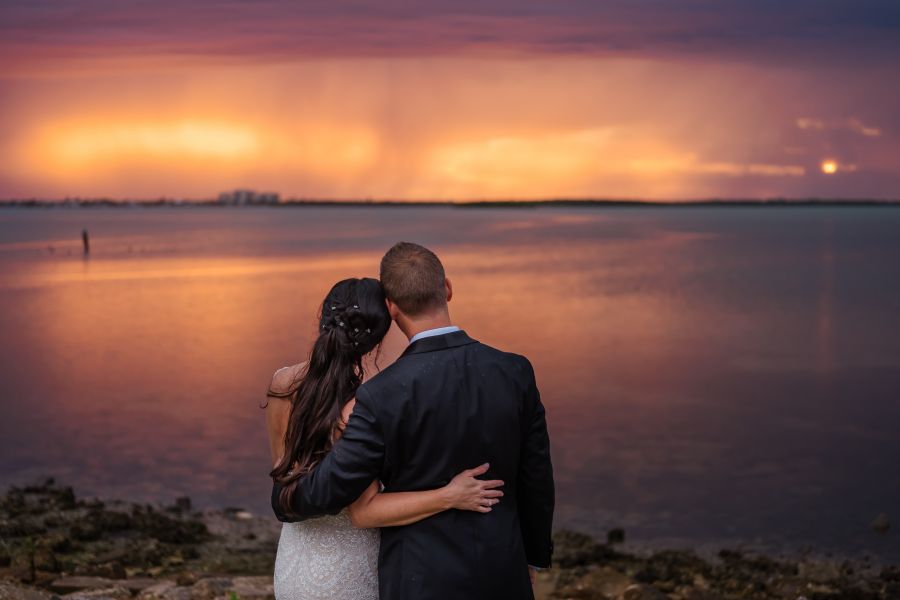 A person in a wedding dress and a person in suit standing on a rocky shore with a sunset in the background